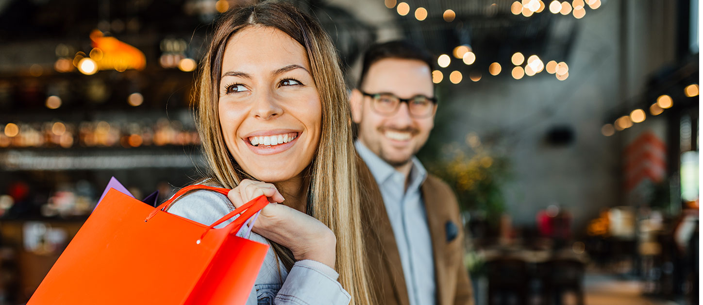 Man and woman smiling while they are shopping. The woman is holding a shopping bag. They are in an area with many things to do.